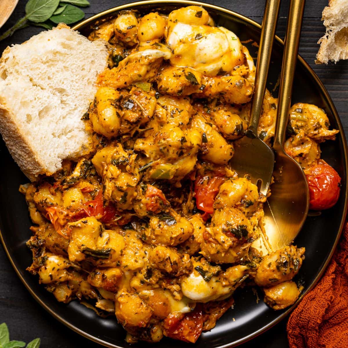 Baked chicken and gnocchi on a plate with a side of bread and two spoons on a black table.