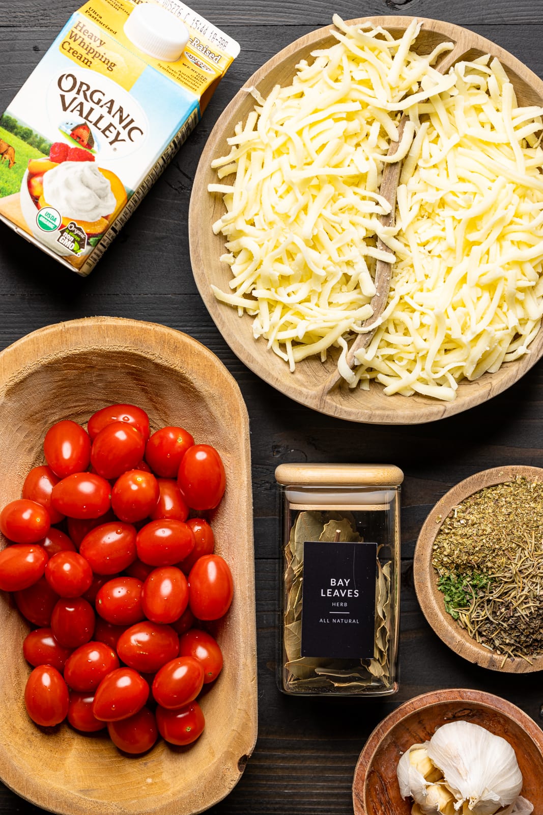 Ingredients on a black table including heavy cream, shredded cheese, tomatoes, herbs and seasonings, and garlic.