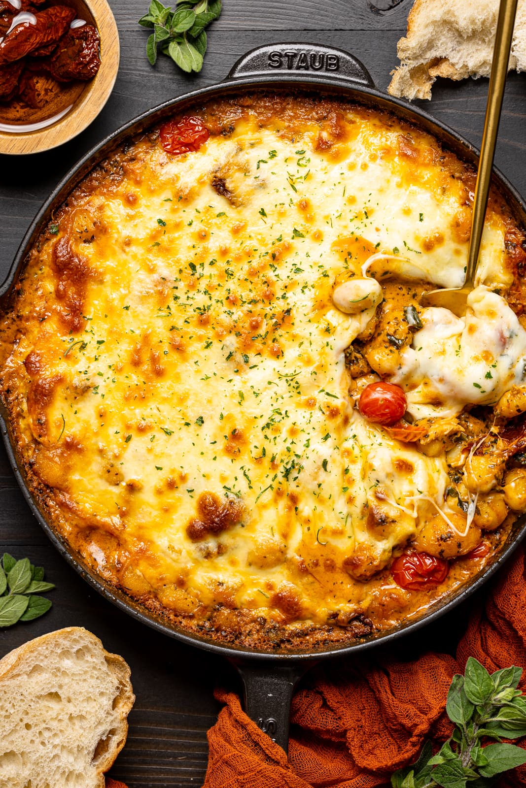 Baked chicken and gnocchi in a skillet, scooped with a spoon on a black table with pieces of bread.