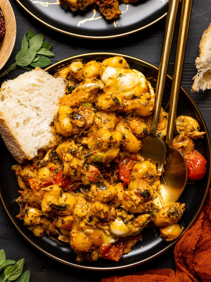 Baked chicken and gnocchi on a plate with a side of bread and two spoons on a black table.