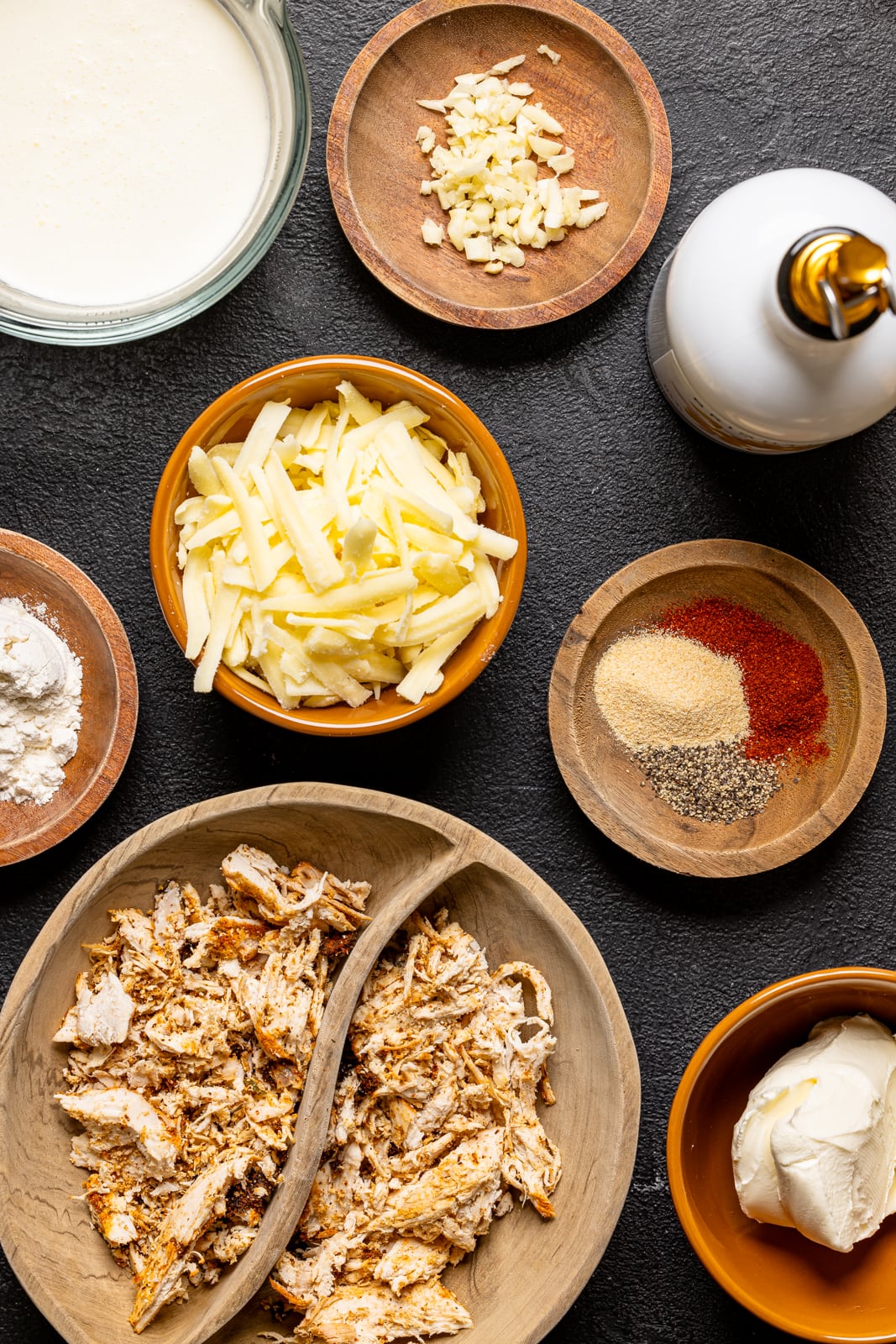 Ingredients on a black wood table including shredded chicken, cheese, garlic, olive oil, flour, heavy cream, cream cheese, and herbs and seasonings. 