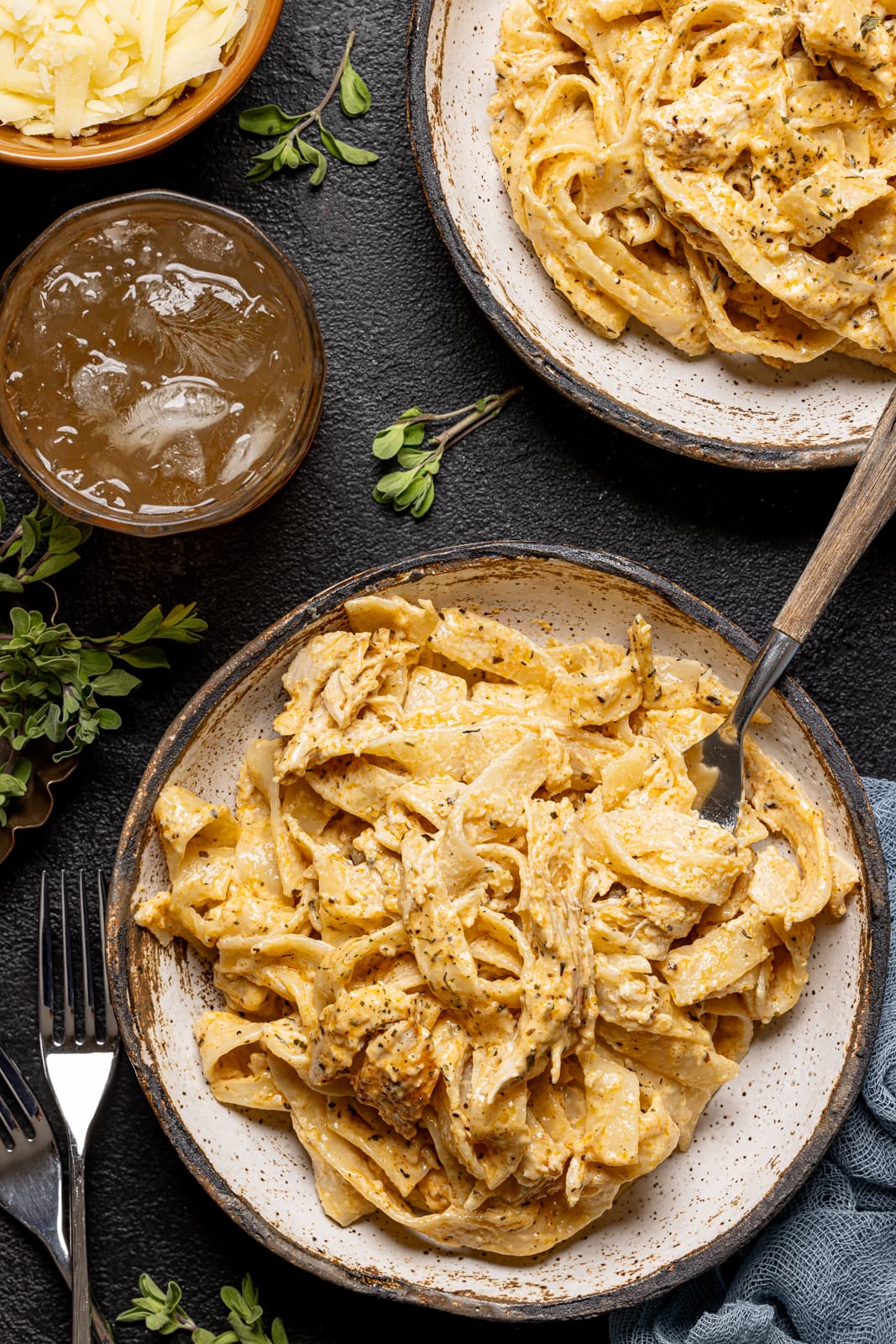 Up close shot of alfredo in two white plates with a drink, side of parmesan cheese, and three forks on a black table.