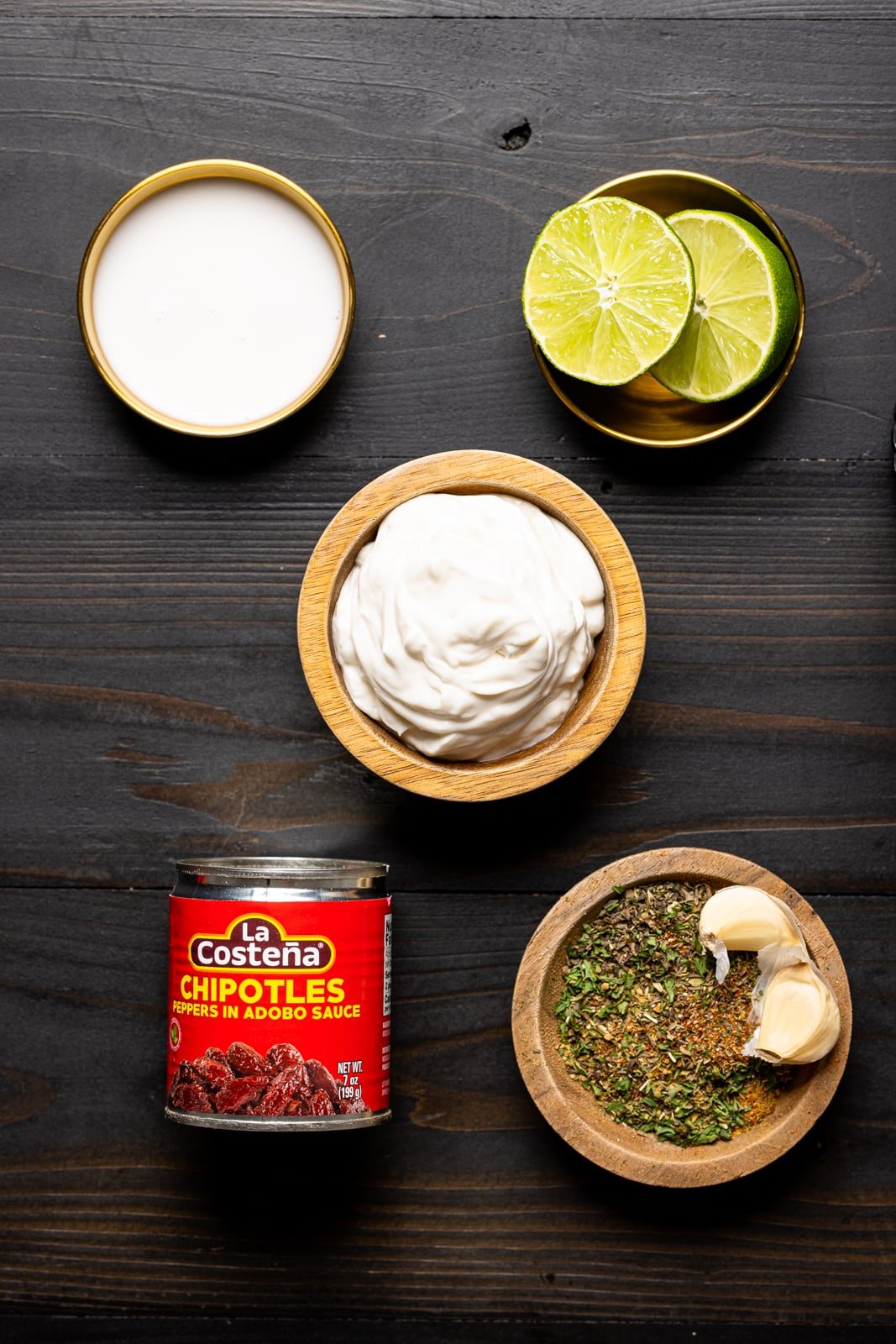 Ingredients on a black wood table including milk, vegan mayonnaise, chipotle peppers, herbs + seasonings, and lime.