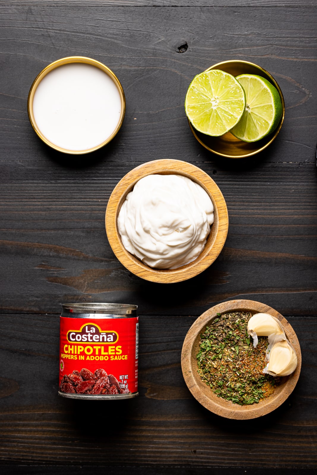 Ingredients on a black wood table including milk, vegan mayonnaise, chipotle peppers, herbs and seasonings, and lime.