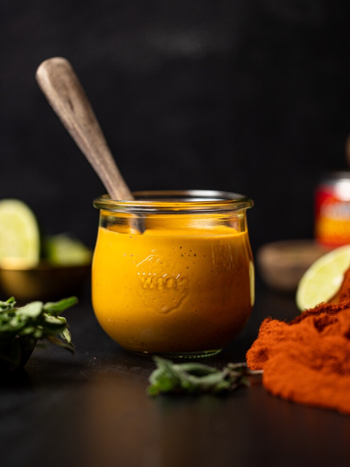 Chipotle sauce in a mason jar with a spoon on a black table with red napkin, lime, and herbs.