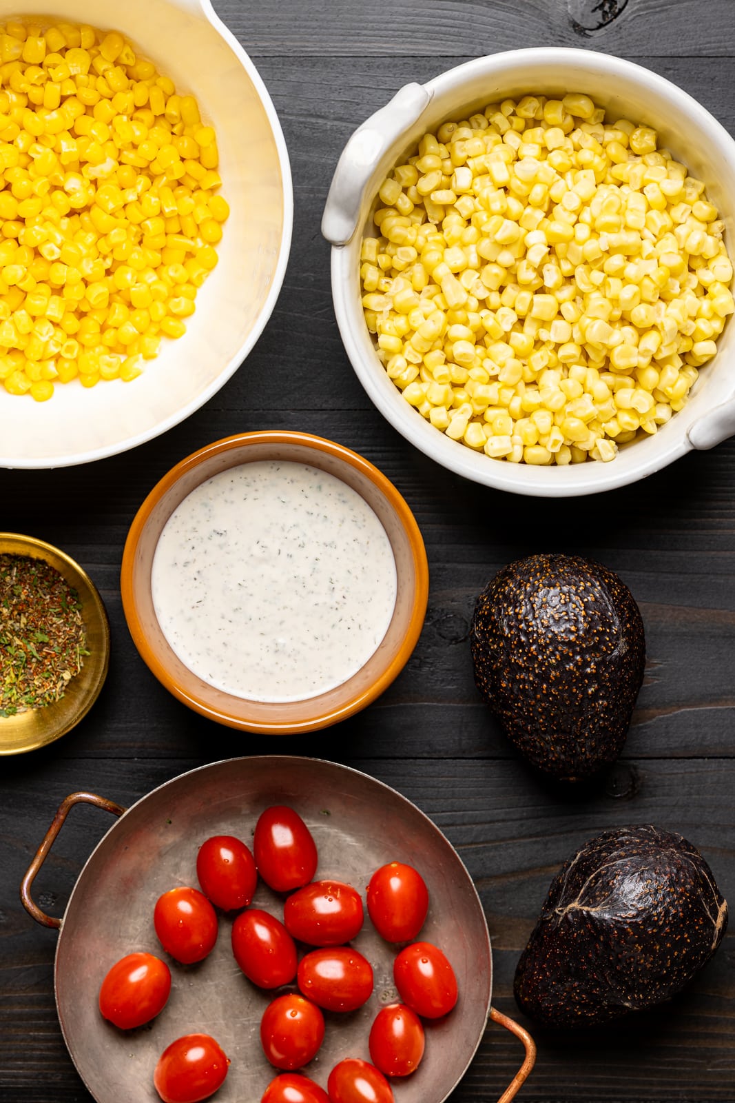 Ingredients on a black wood table including corn, avocado, tomatoes, sauce, and herbs and seasonings.