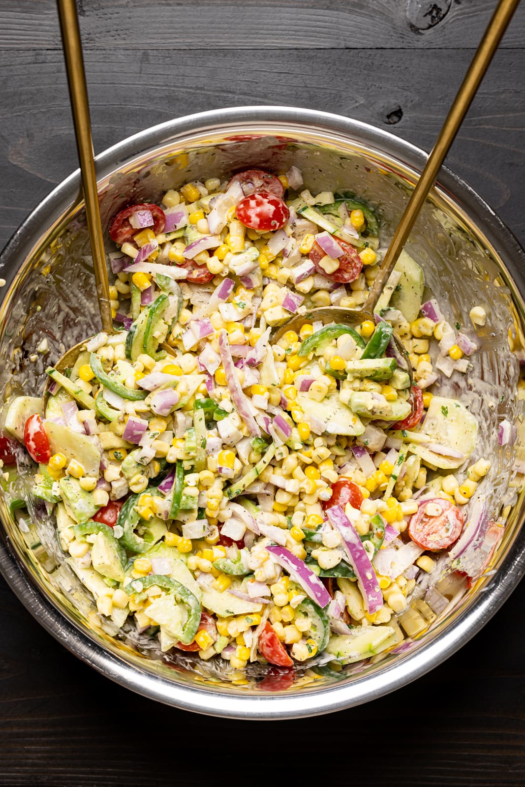 Corn salad ingredients in a large silver bowl with two spoons on a black wood table.