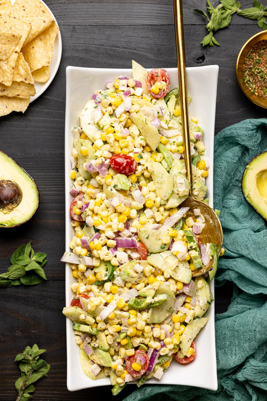 Long white plate of salad with a spoon on a black wood table with tortilla chips and sliced avocados.