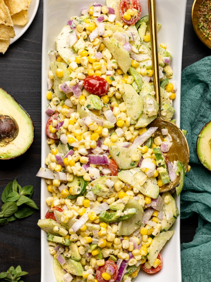 Salad in a long white plate with a spoon on a black wood table with tortilla chips and sliced avocados.