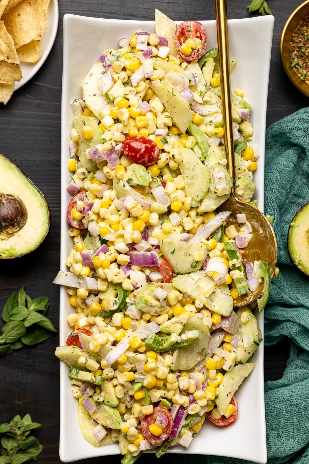 Salad in a long white plate with a spoon on a black wood table with tortilla chips and sliced avocados.