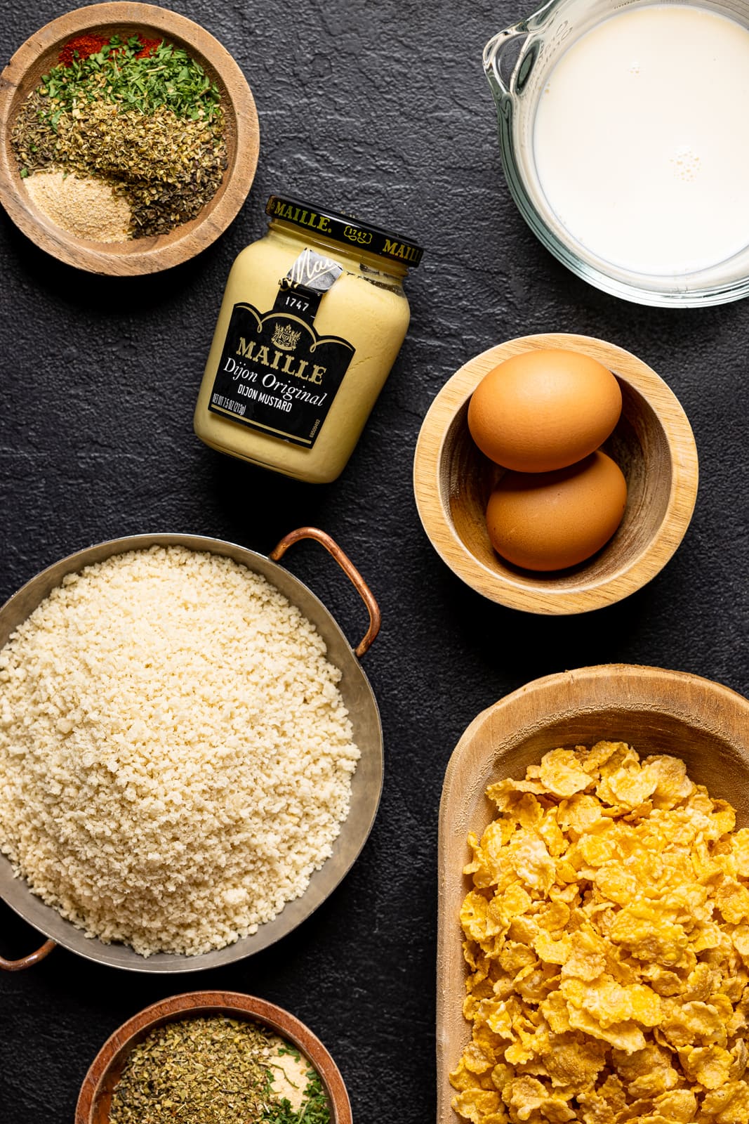 Ingredients on a black table including cornflakes, breadcrumbs, seasonings, eggs, dijon mustard, and milk.