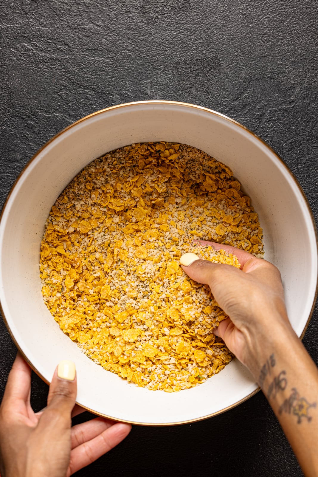 Crushed cornflakes in a white bowl with hands mixing it around.