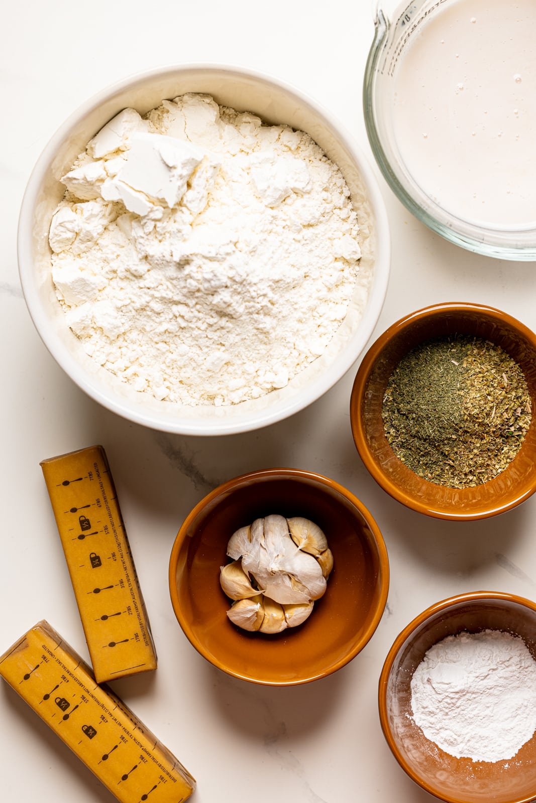 Ingredients on a white table including flour, butter, garlic, herbs + seasonings, milk, and baking powder.