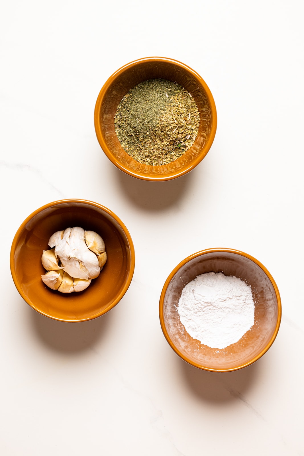Garlic, herbs + seasonings, and baking powder in brown pinch bowls on a marble table.