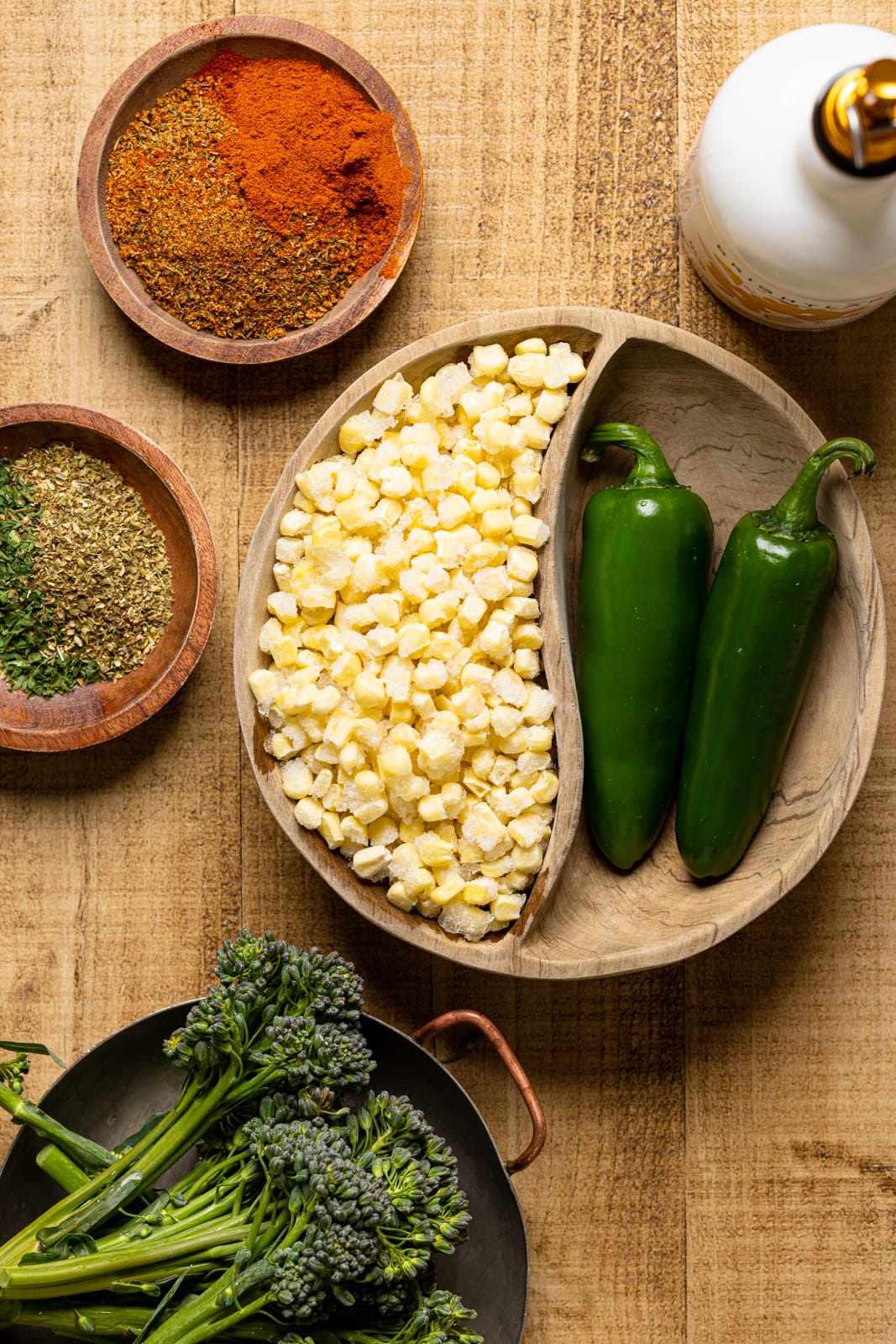 Ingredients of salad on a brown wood table including broccolini, jalapeños, corn, seasonings and herbs.