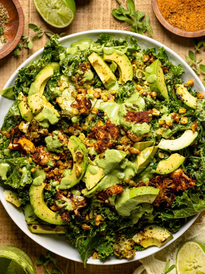 Salad on a white round plate with seasonings and herbs, lime wedges, dressing, and a yellow napkin.