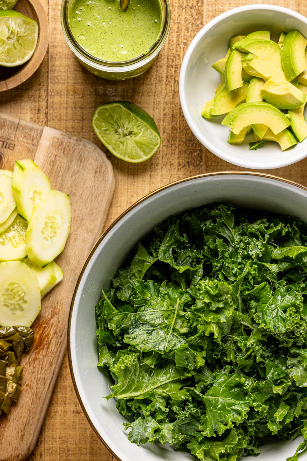 Ingredients on a wood table including kale, cucumber, lime, dressing, and avocado.