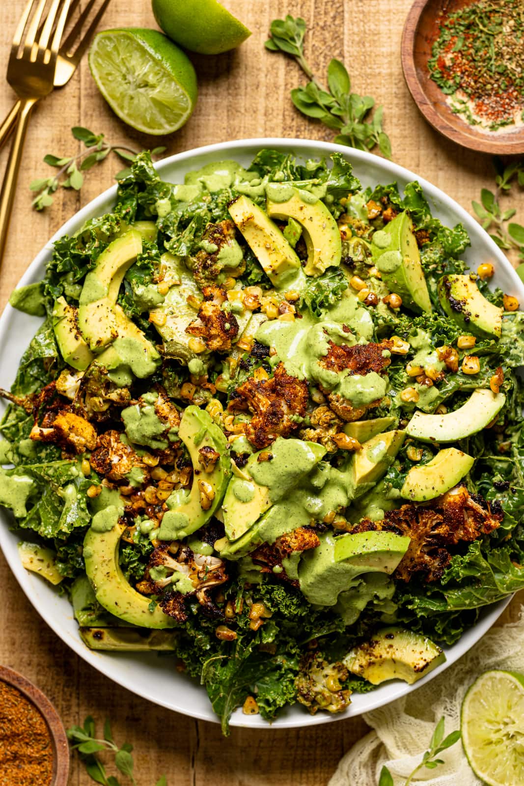 Salad on a white round plate on a wood table with two gold forks, lime wedges, and seasonings and herbs.