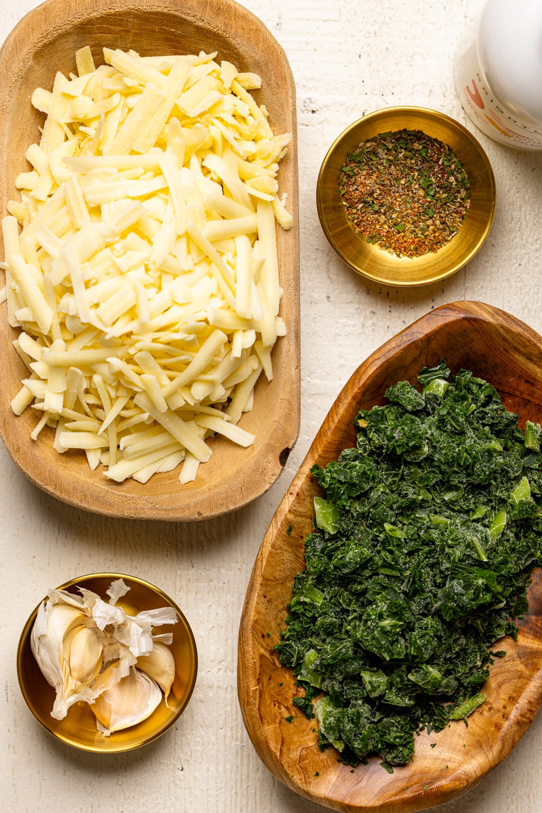 Ingredients on a white wood table including chopped kale, garlic, cheese, and herbs + seasonings. 