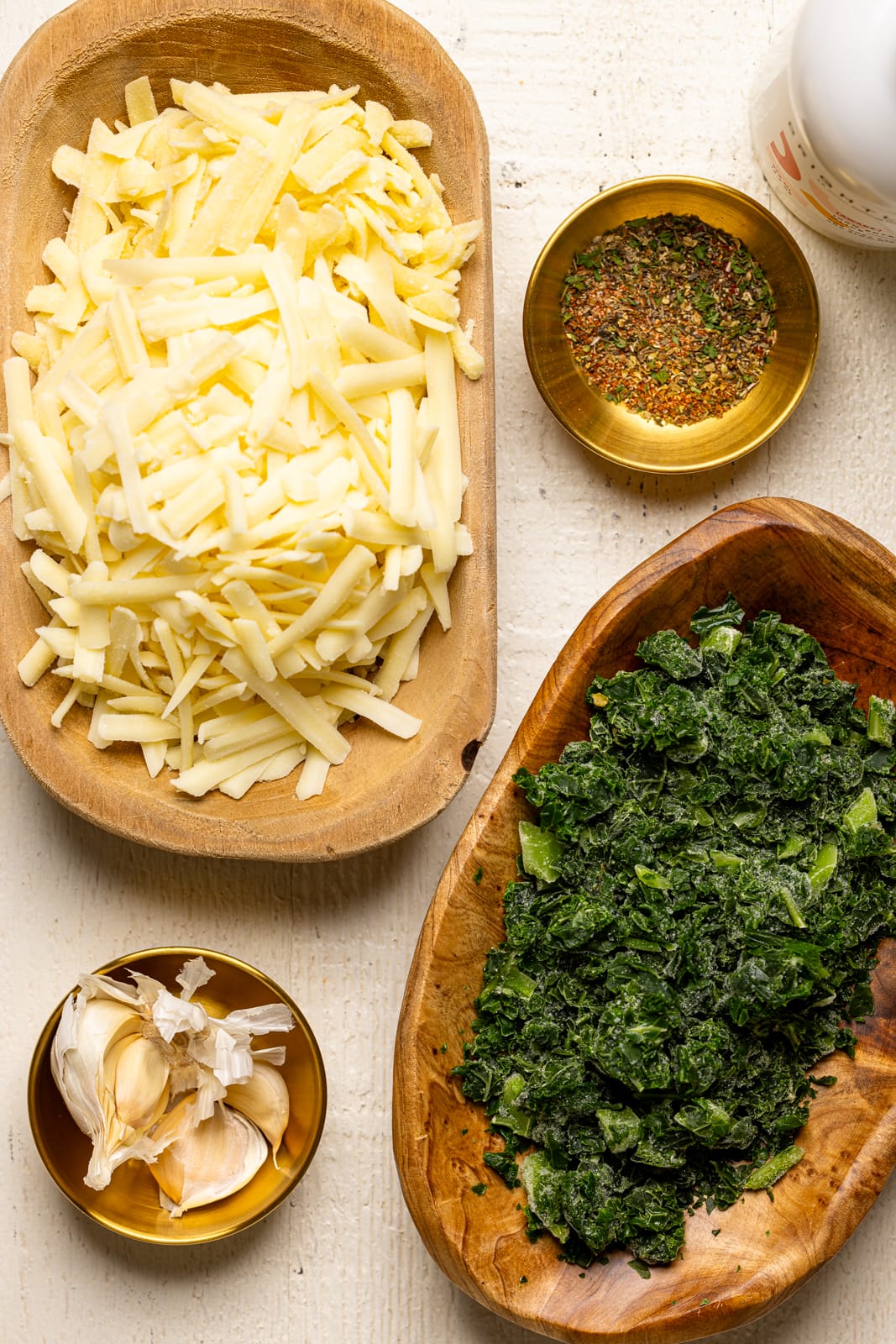 Ingredients on a white wood table including chopped kale, garlic, cheese, and herbs and seasonings. 