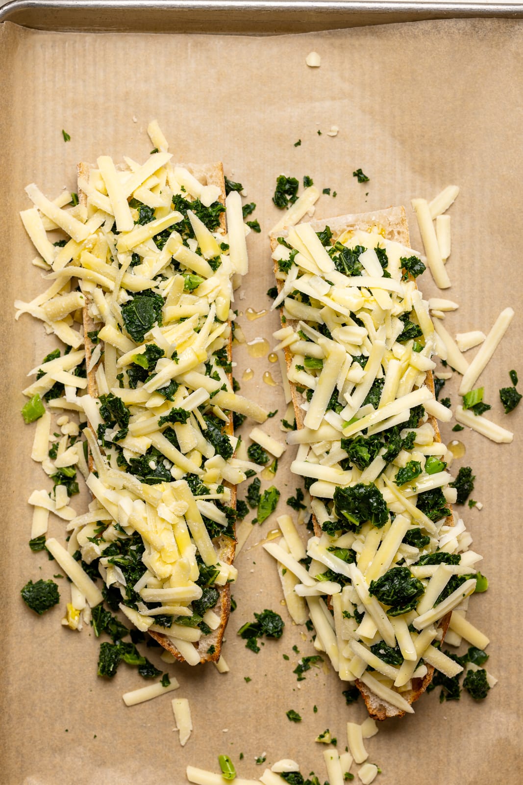 Two slices of french bread on a baking sheet with parchment paper with cheese and chopped kale.