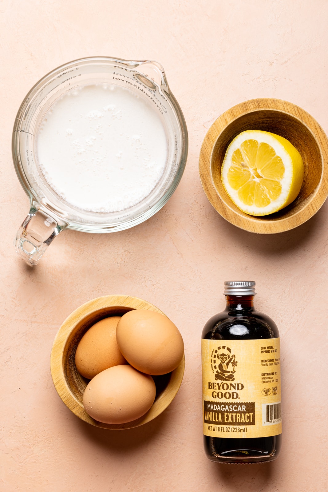 Ingredients on a peach colored table including milk, vanilla, eggs, and lemon.