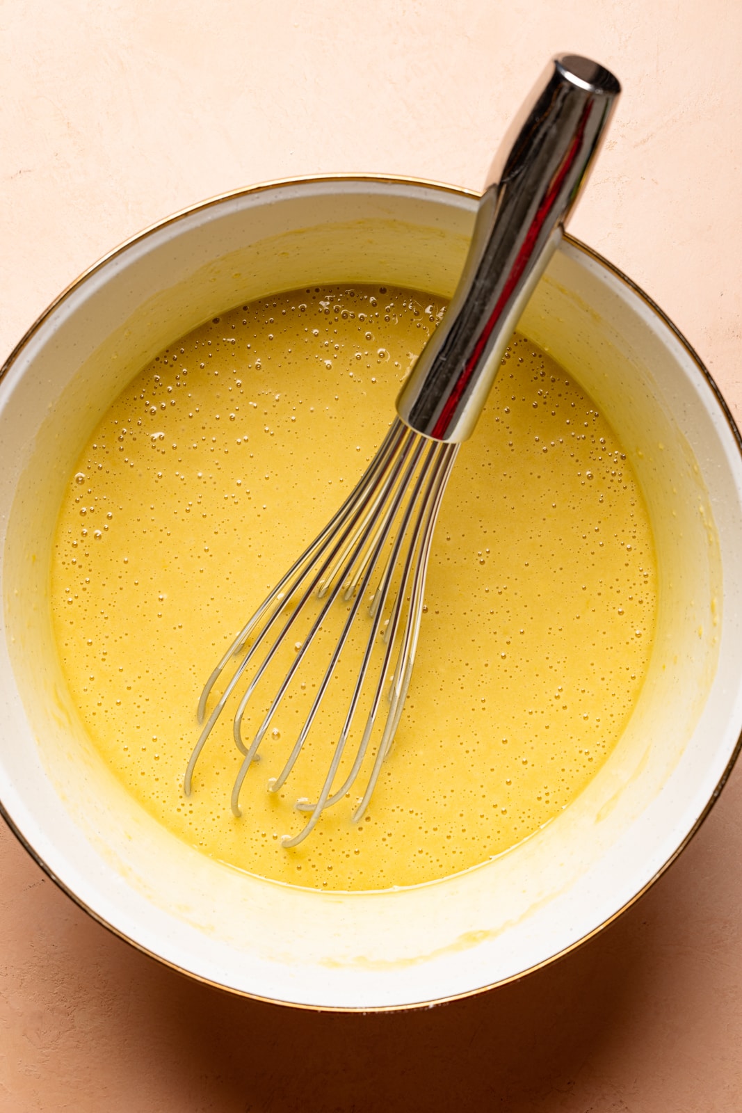 Cake batter in a large white bowl with a whisk on a peach colored table.