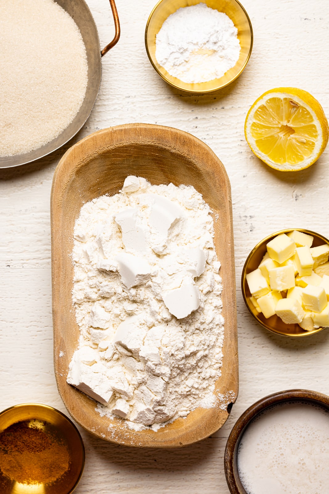 Ingredients on a white wood table including flour, sugar, baking powder, spices, butter, milk, and lemon.