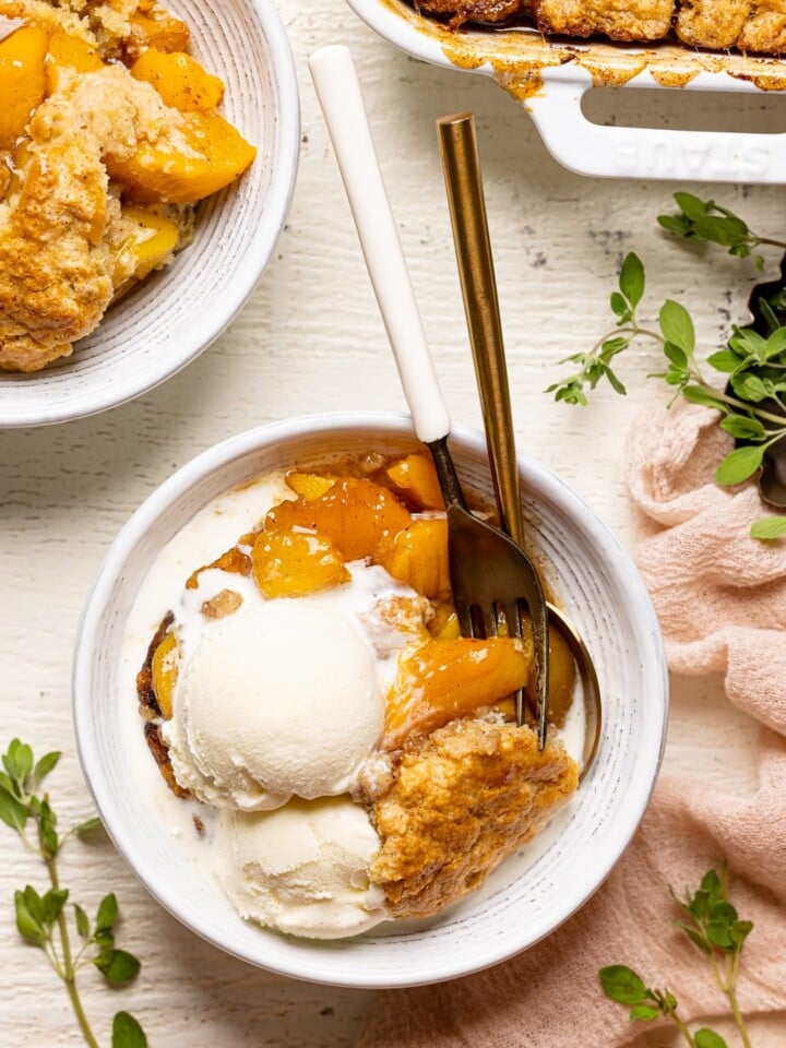 Peach cobbler in a white bowl on a white wood table next to a plate and baking dish and herbs.