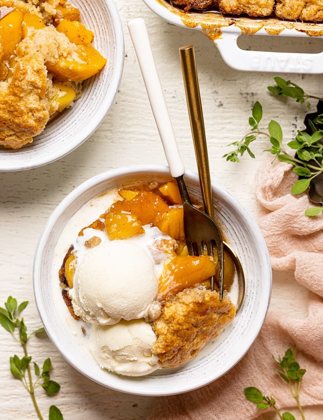White bowl of peach cobbler on a white wood table next to a plate and baking dish and herbs.