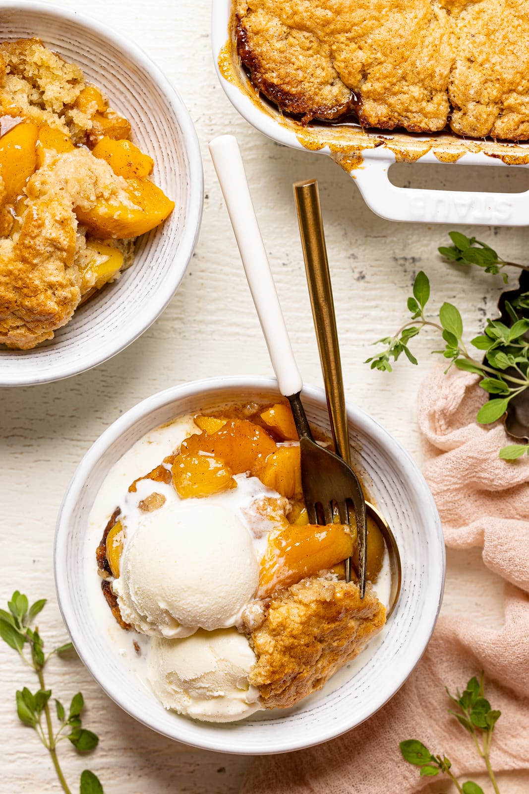 White bowl of peach cobbler on a white wood table next to a plate and baking dish and herbs.