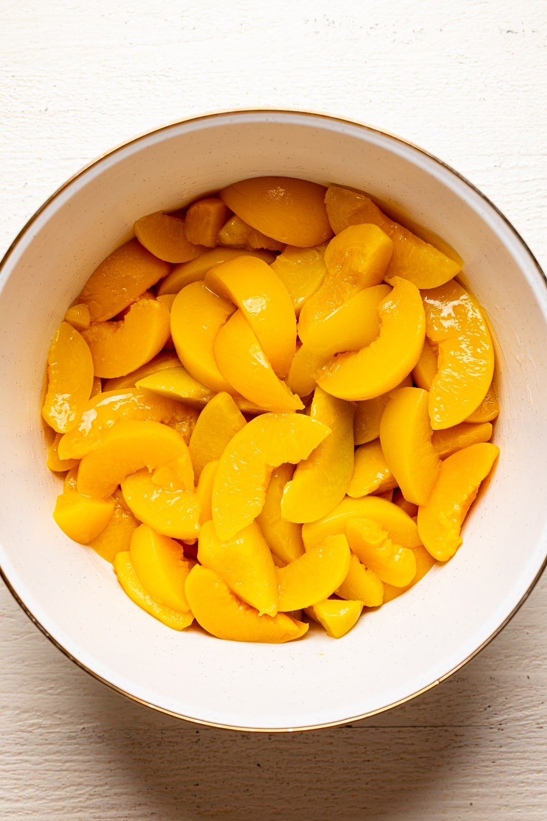 Peach slices in a large white bowl on a white wood table.
