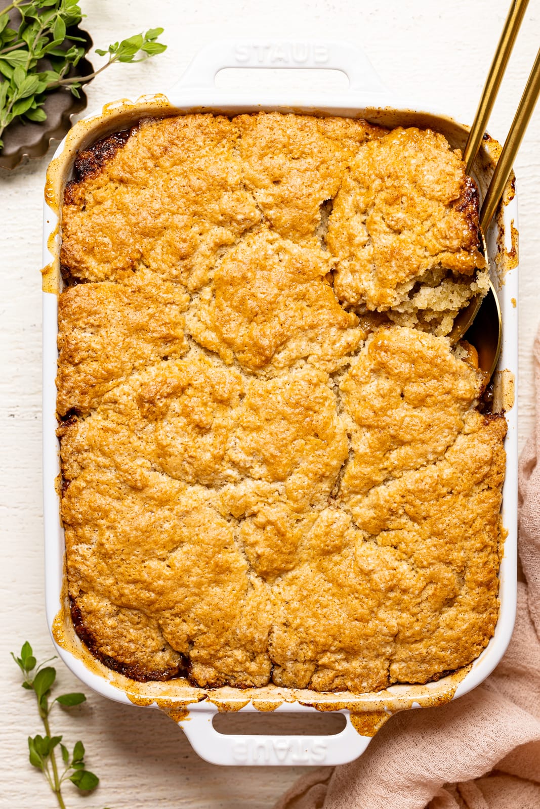 Baked peach cobbler in a white baking dish with two serving spoons on a white wood table with a pink napkin.