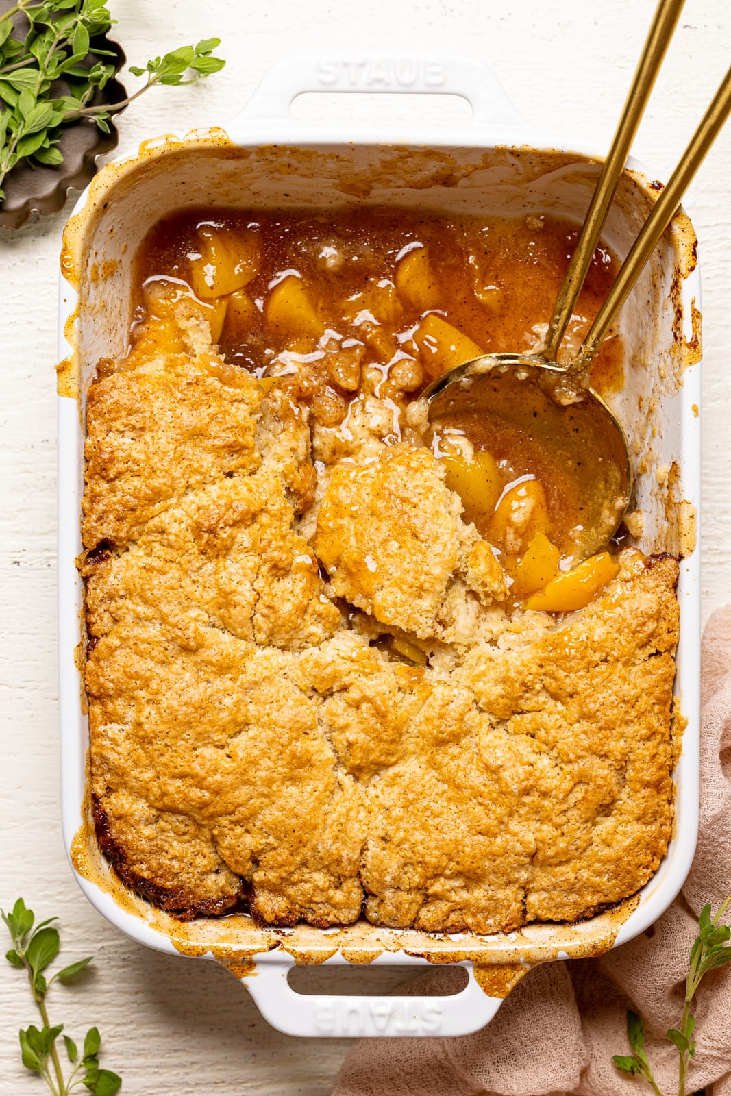 Peach cobbler scooped out in a white baking dish with two serving spoons on a white wood table with a pink napkin.