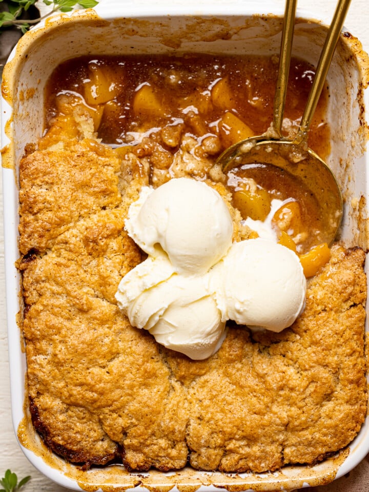 Up close shot of peach cobbler in baking dish with scoops of ice cream on top and two serving spoons.