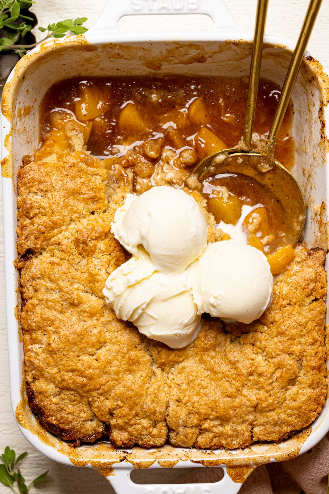 Up close shot of peach cobbler in baking dish with scoops of ice cream on top and two serving spoons.