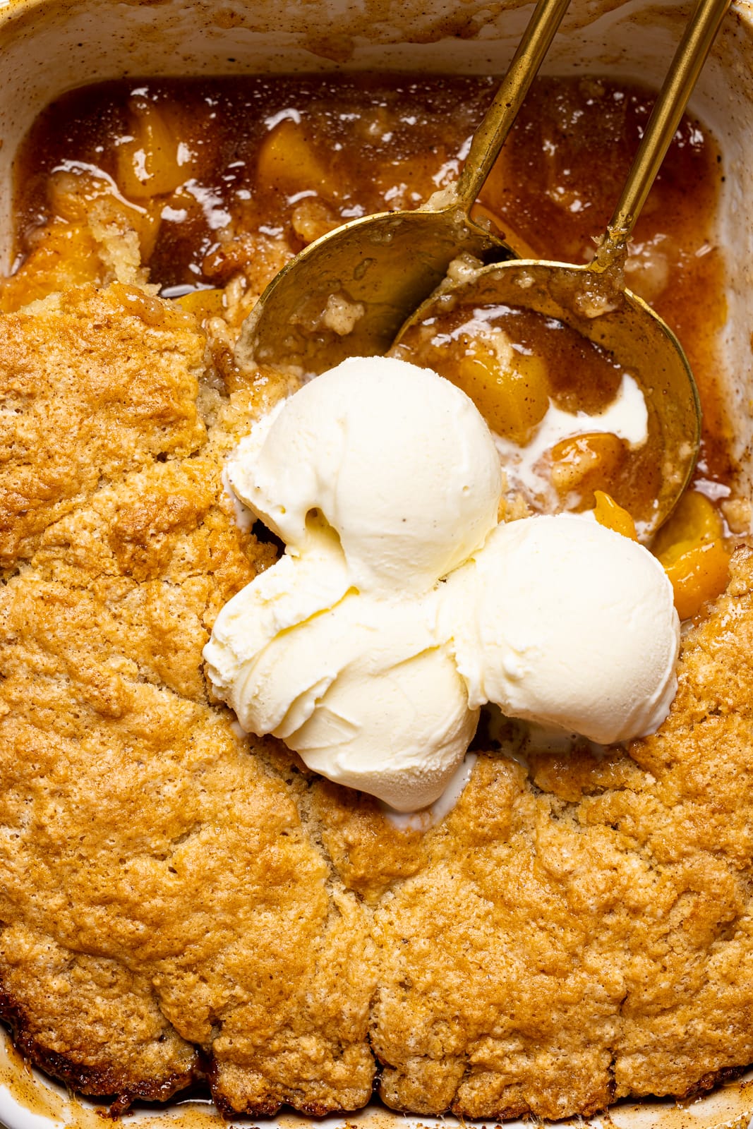 Very up close shot of peach cobbler in baking dish with scoops of ice cream on top and two serving spoons.