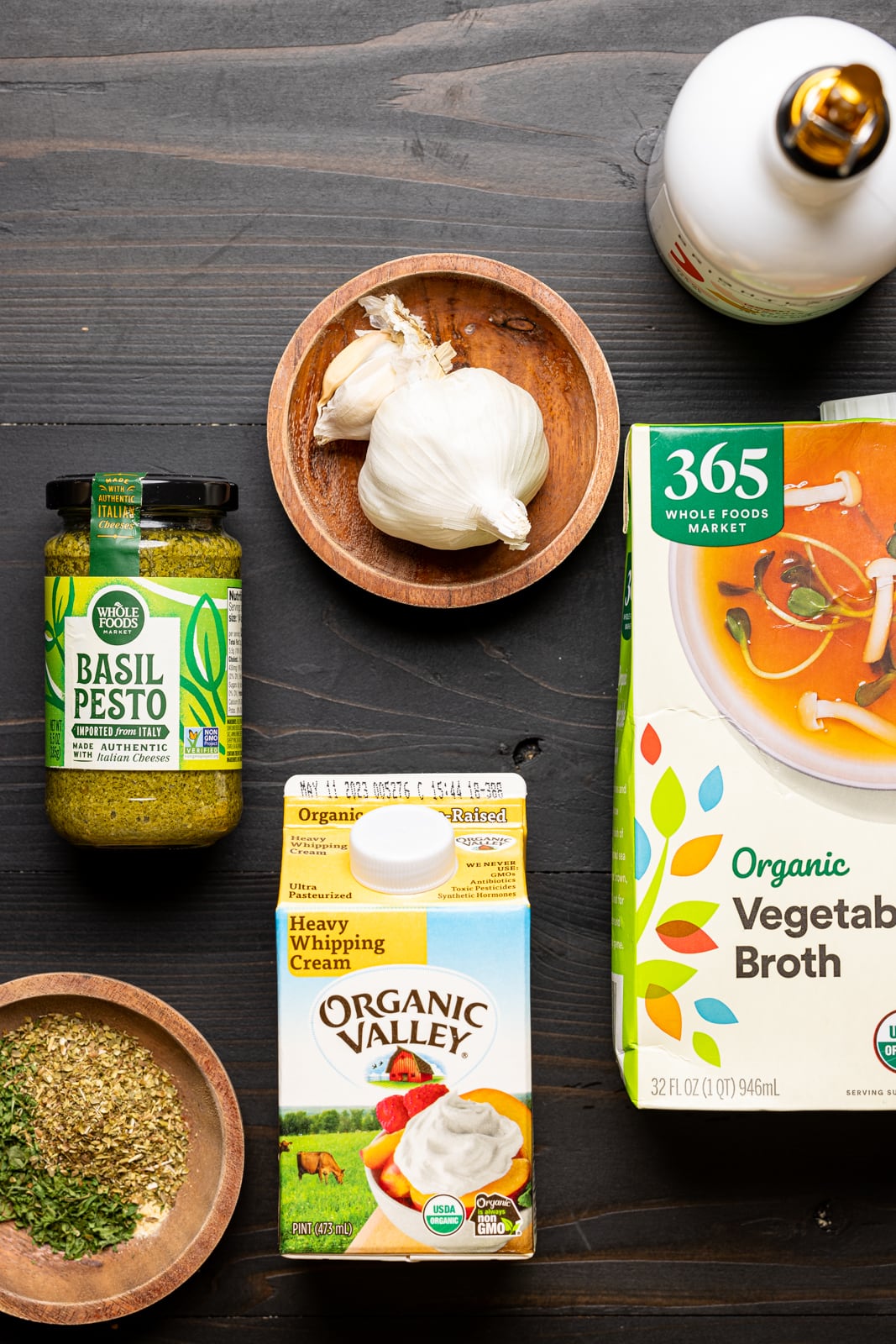 Ingredients on a black table including garlic cloves, olive oil, pesto sauce, heavy cream, vegetable stock, and seasonings.