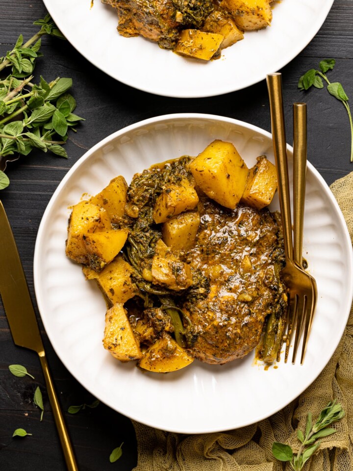 Chicken and potatoes on two white plates on a black table with forks and knives and herbs.