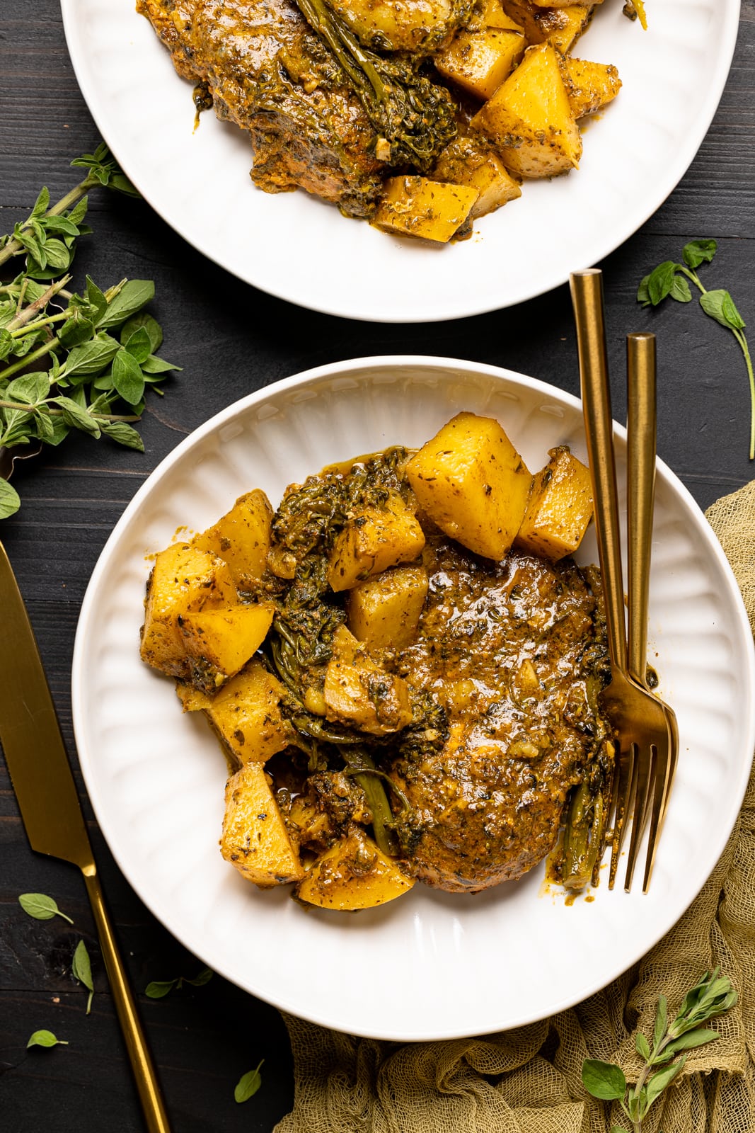 Chicken and potatoes on two white plates on a black table with forks and knives and herbs.