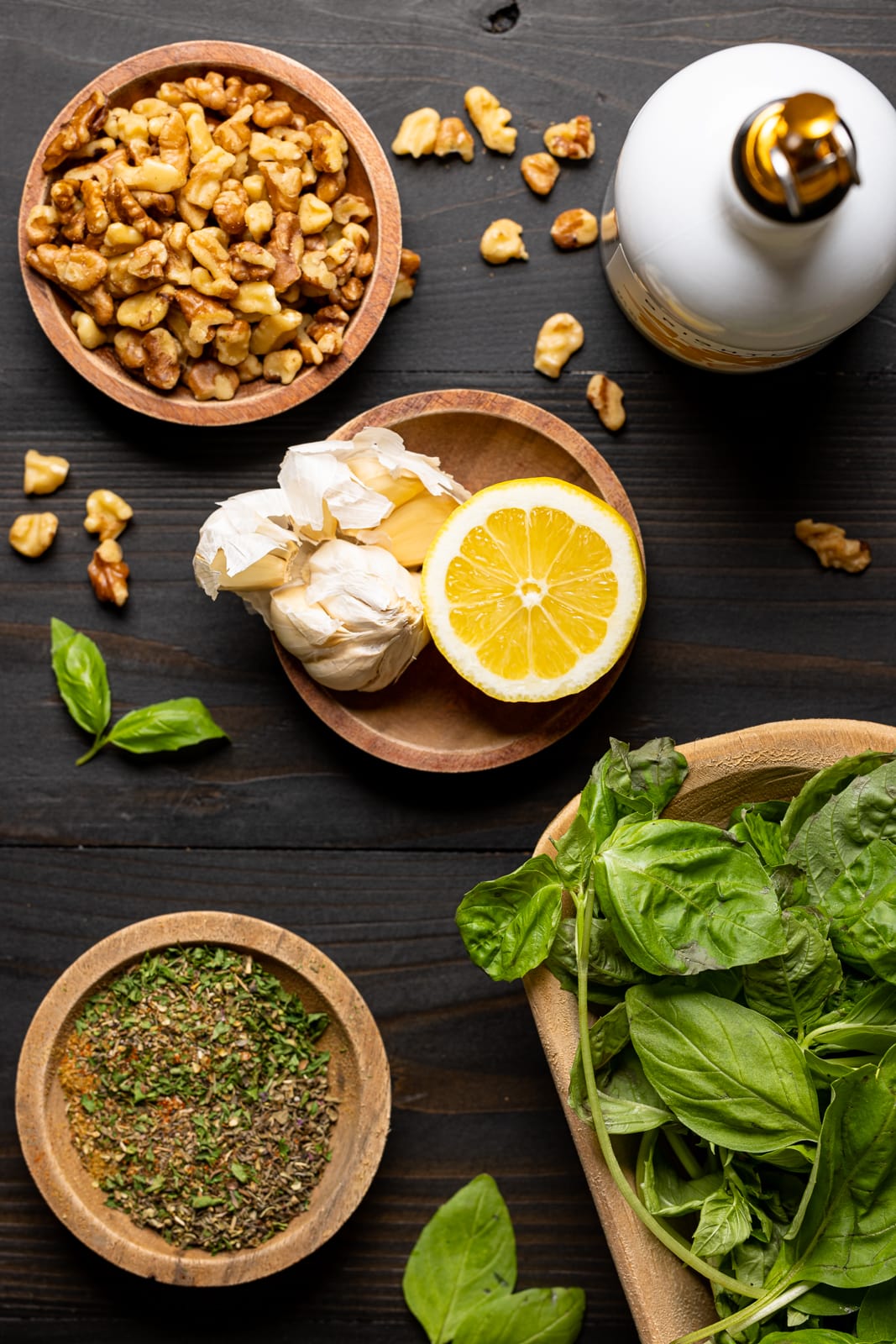 Ingredients on a black wood table including walnuts, olive oil, lemon, garlic cloves, herbs and seasonings, and fresh basil.