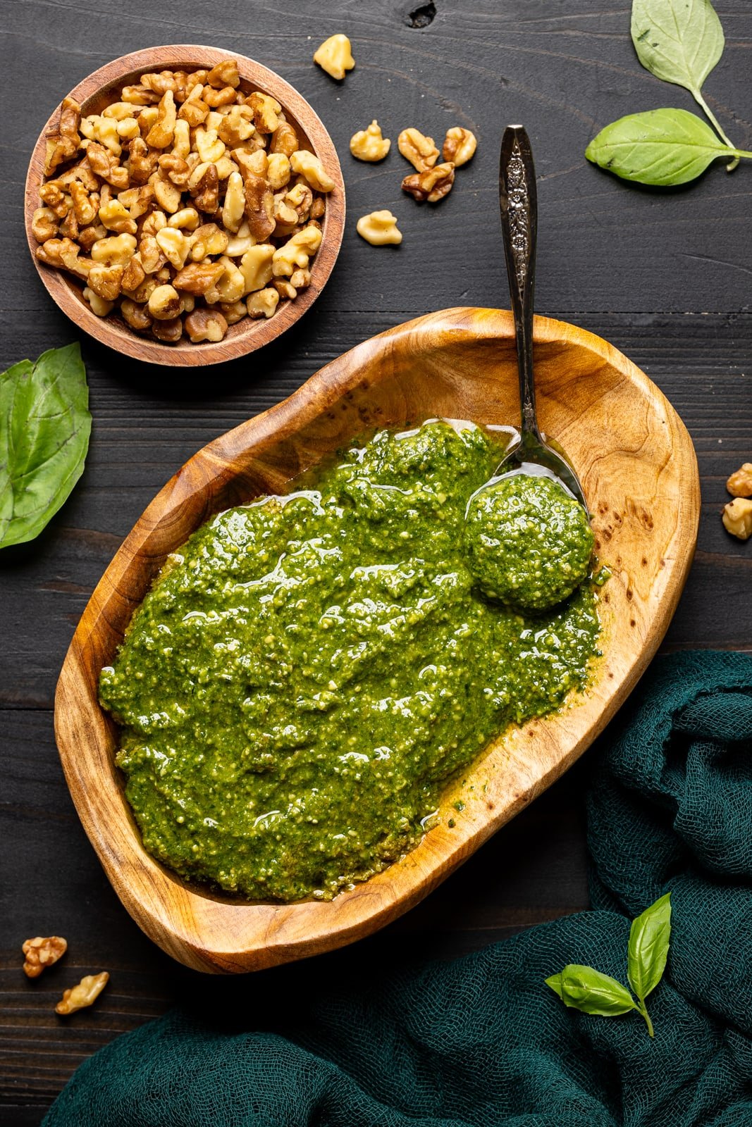 Pesto sauce in a brown wood bowl on a black wood table with walnuts and basil leaves.