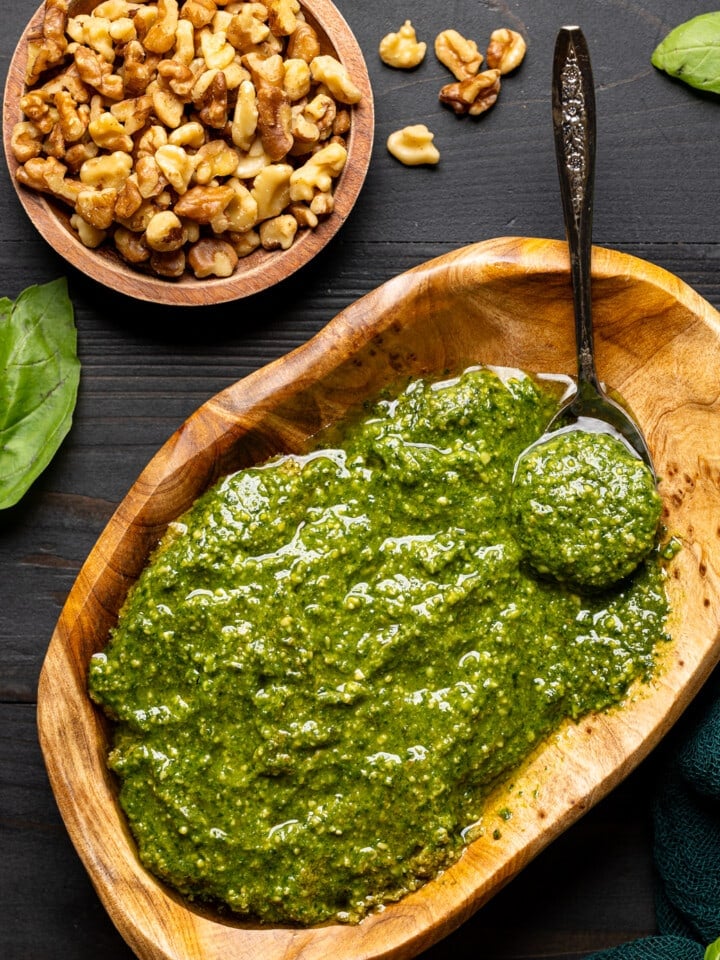 Pesto sauce in a brown wood bowl with a spoon on a black wood table with walnuts and basil leaves.