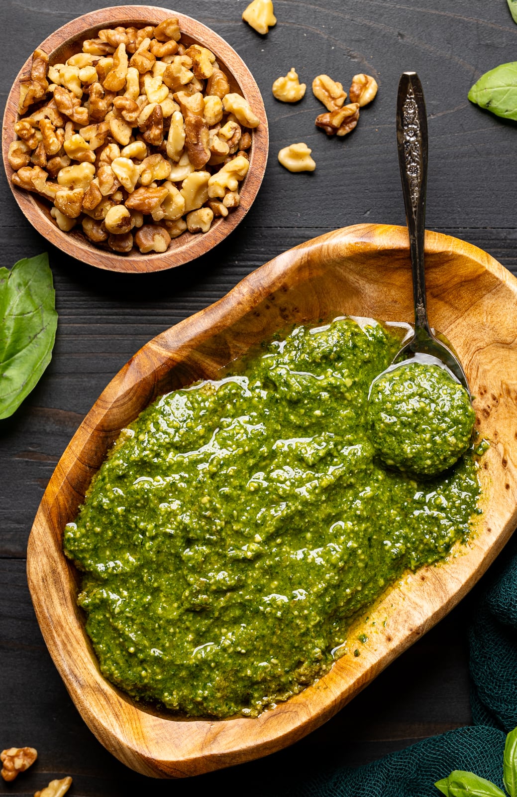 Pesto sauce in a brown wood bowl with a spoon on a black wood table with walnuts and basil leaves.