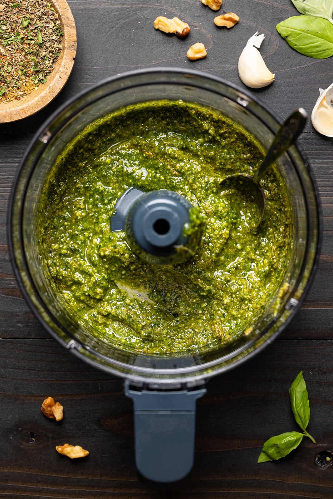 Sauce in a food processor with a spoon and garnish of walnuts, garlic, herbs and seasonings, and basil leaves.