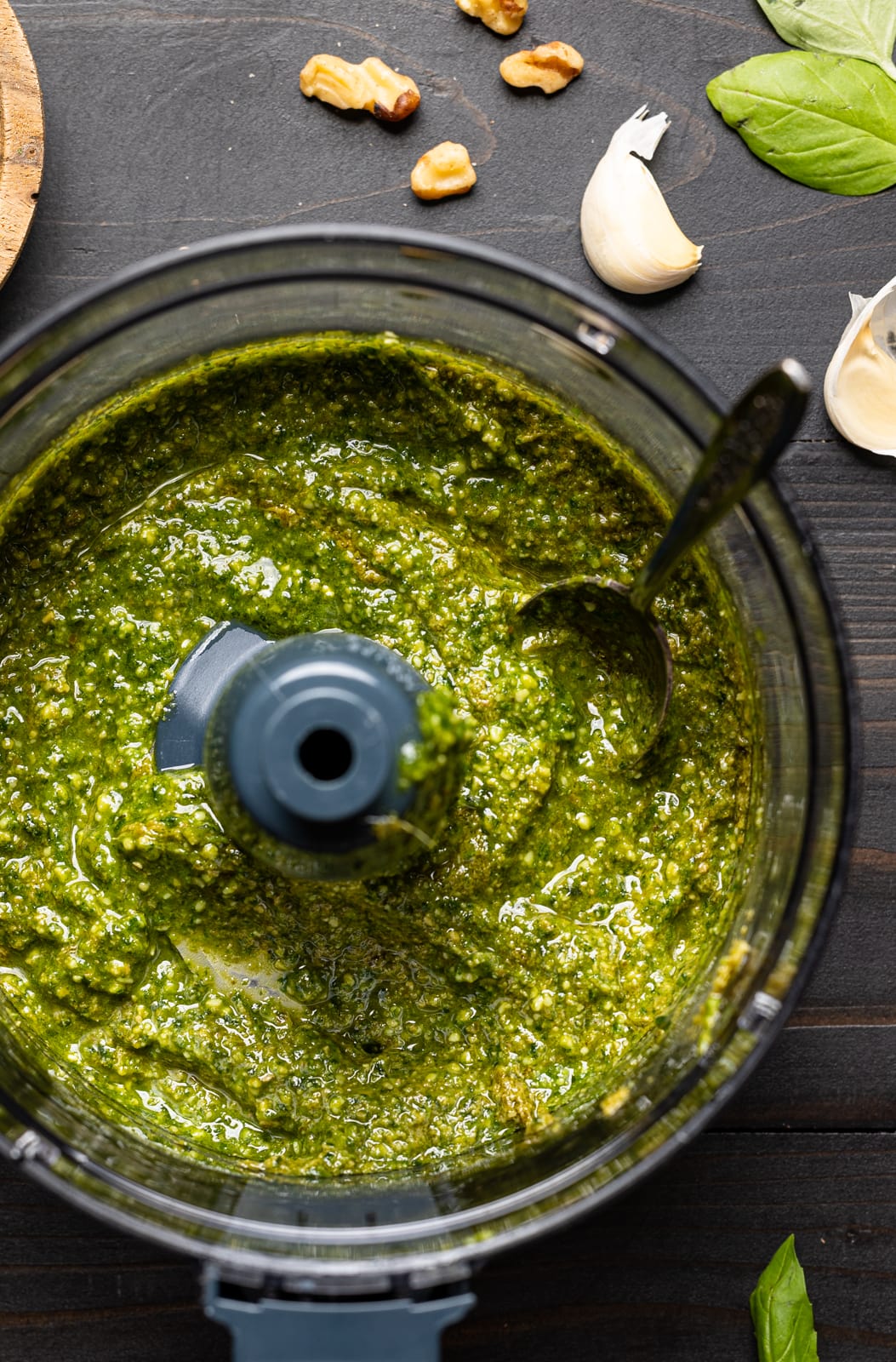 Pesto sauce in a food processor on a black wood table with garnishes of walnuts and garlic cloves.
