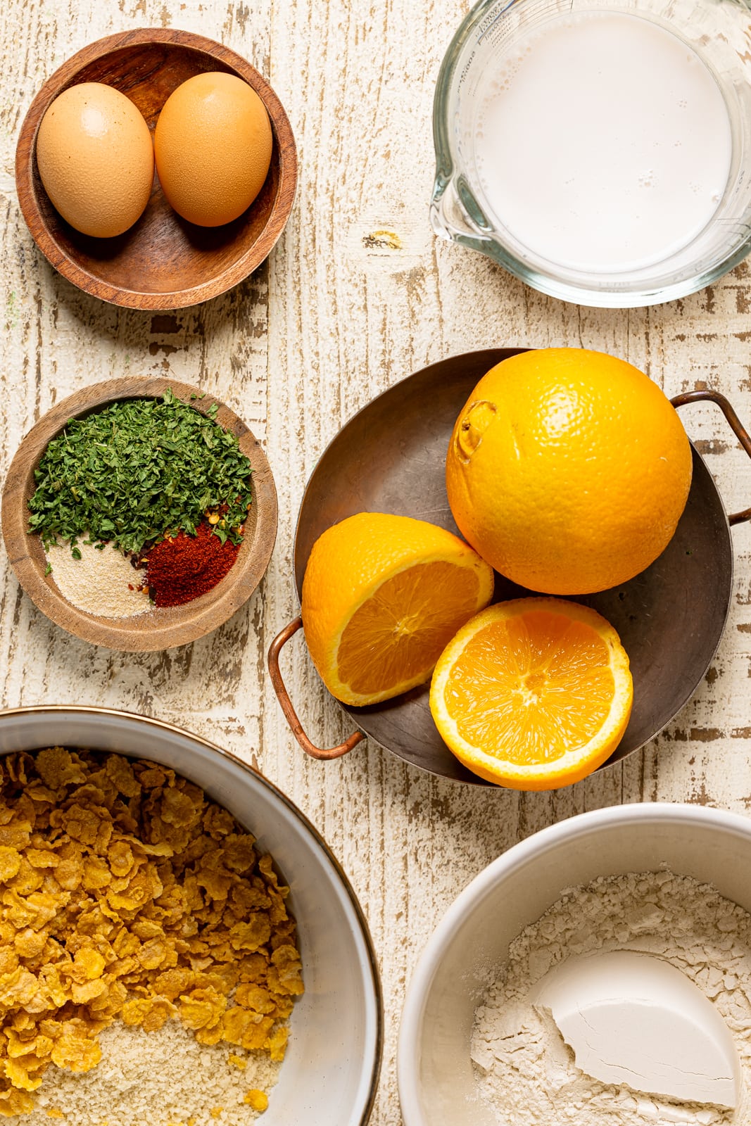 Ingredients on a white wood table including eggs, herbs and seasonings, milk, oranges, cornflakes, and flour. 