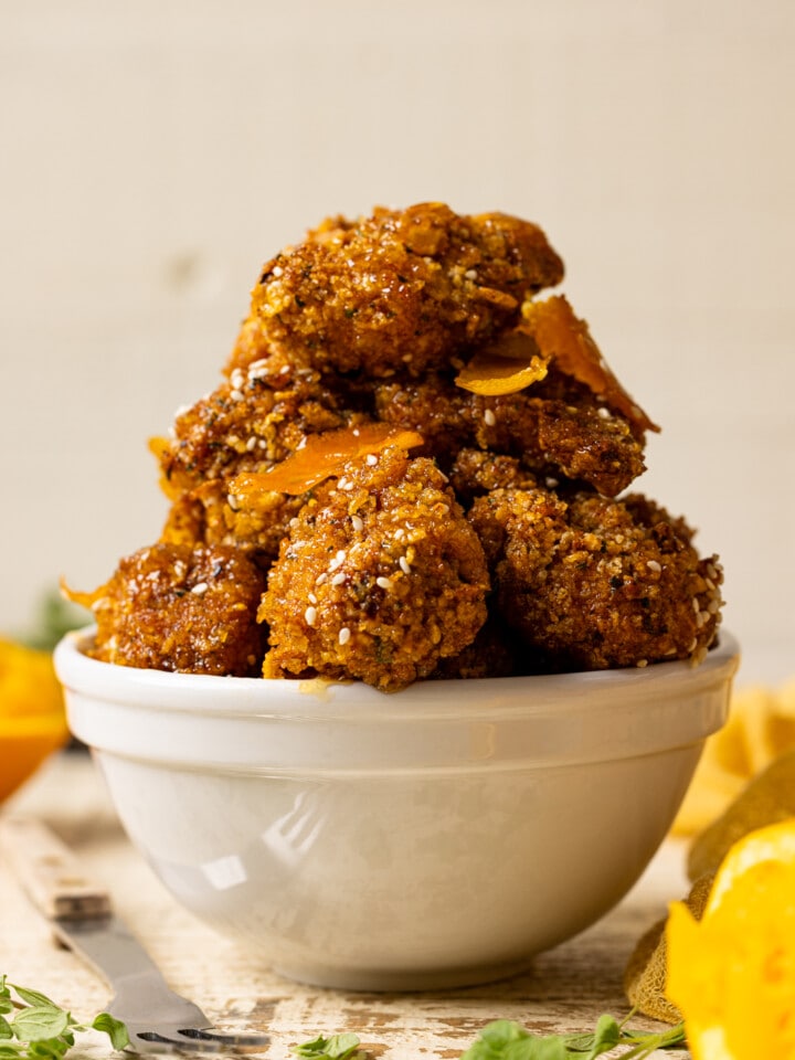 Up close shot of chicken stacked in a white bowl on a white wood table with oranges and herbs.