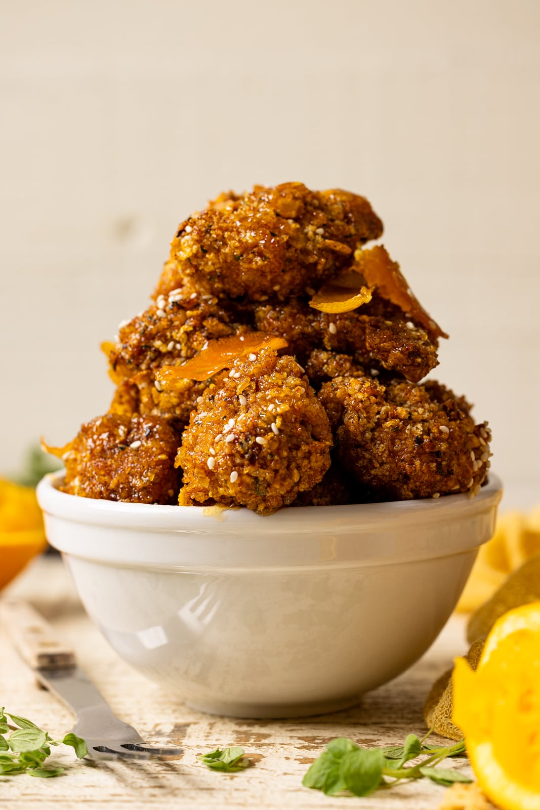 Up close shot of chicken stacked in a white bowl on a white wood table with oranges and herbs.