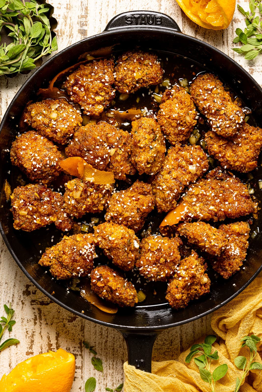 Popcorn chicken in a black skillet on a white wood table with oranges and herbs.